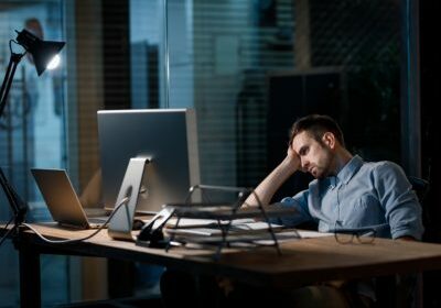 Casual man looking fatigue while working with computer in dark office alone.
